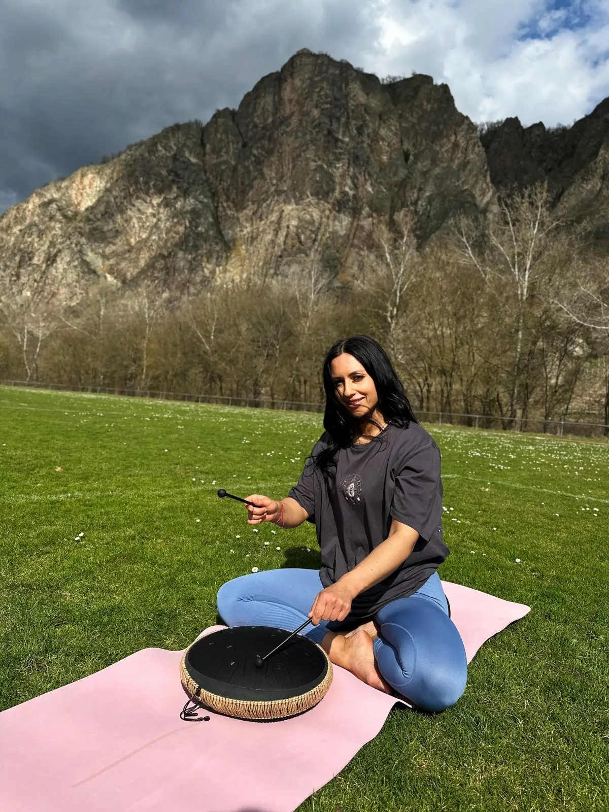 Mascha spielt eine Rahmentrommel auf der Matte, über dem Fels ein dramatischer Himmel